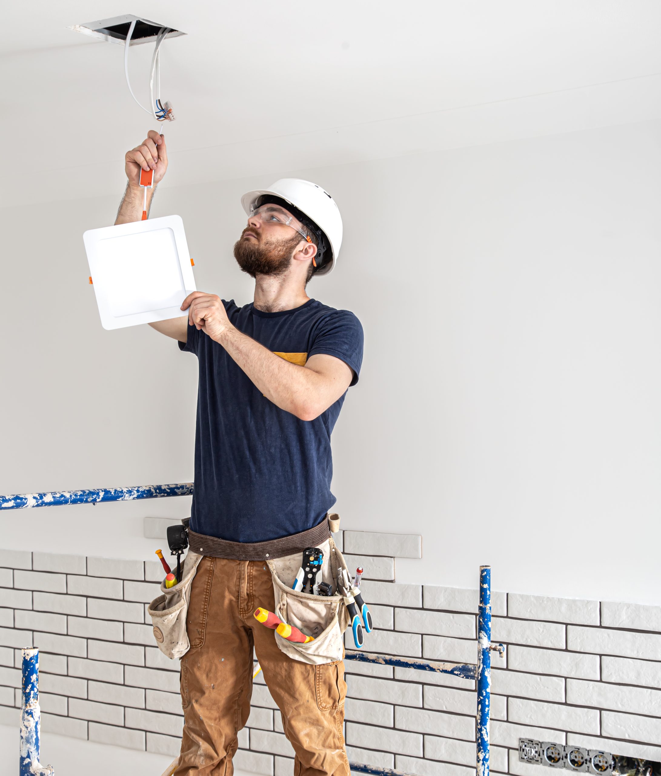 Electrician Builder with beard worker in a white helmet at work, installation of lamps at height. Professional in overalls with a drill on the background of the repair site.